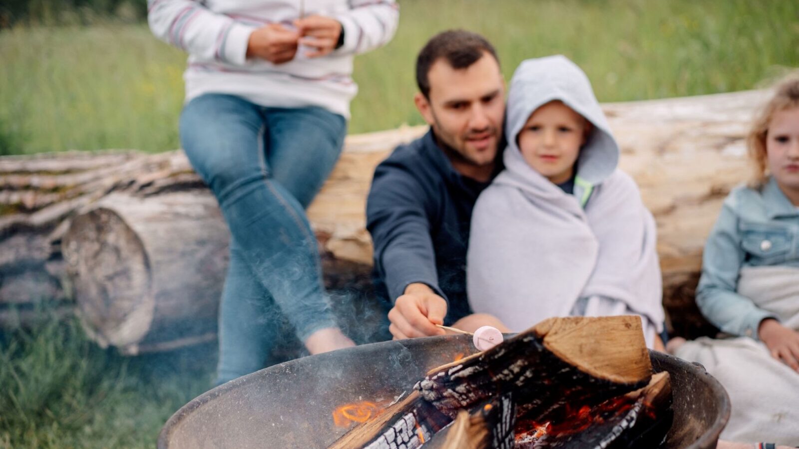 A family sits around a crackling campfire, embracing the serenity of Nature’s Nest. The man holds a marshmallow on a stick over the dancing flames, while a young child wrapped in a blanket snuggles on his lap. A log serves as their bench amidst the lush grass, perfectly capturing the essence of glamping.