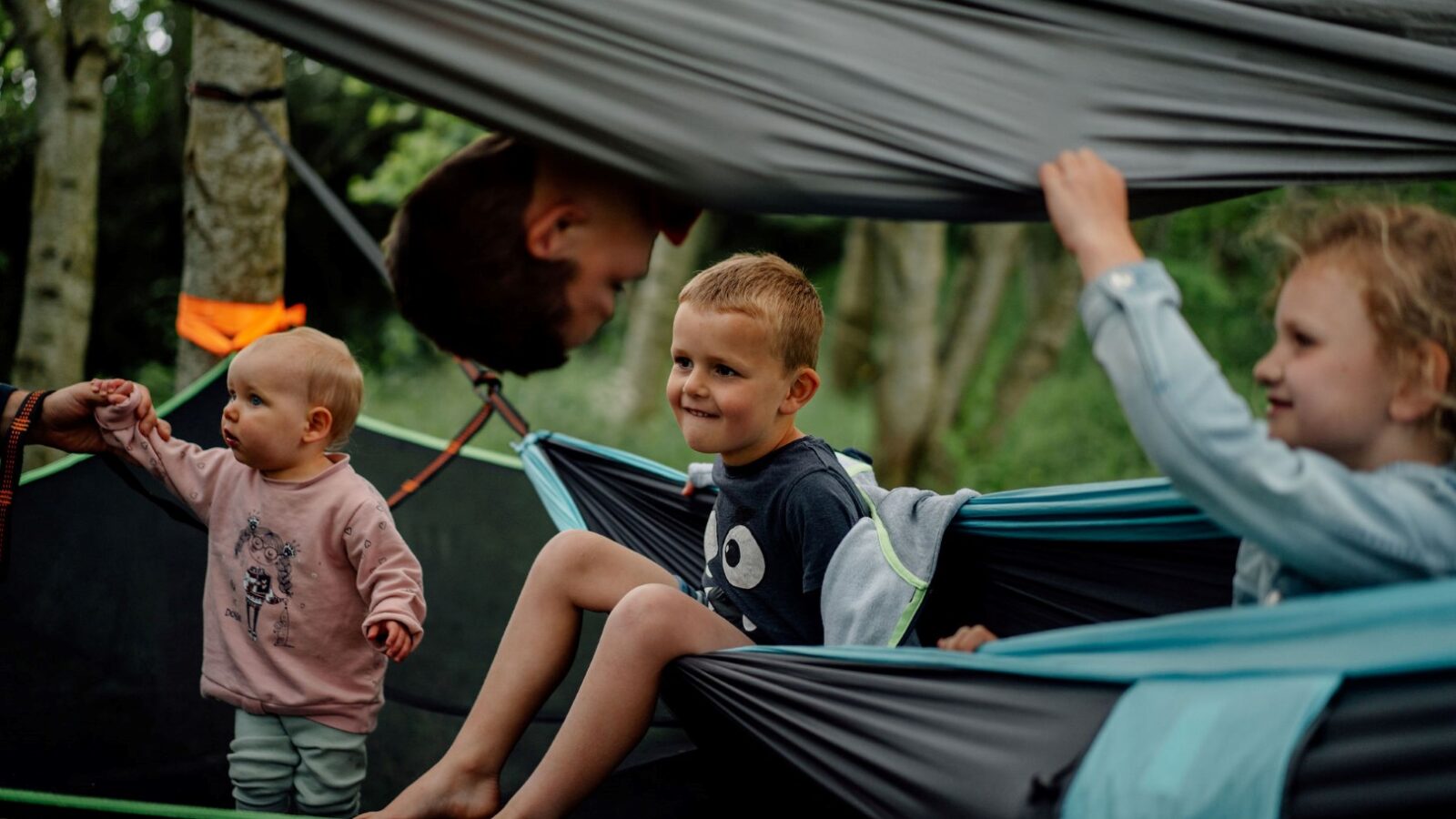 Three children are playing in hammocks suspended between trees, surrounded by the tranquility of nature. Nearby, a baby stands holding onto a hammock, adding to the lively scene in this outdoor nest on a cloudy day.