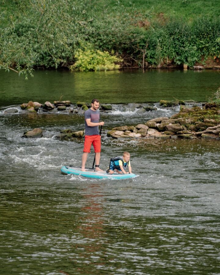 A man in a black shirt and red shorts stands on a paddleboard at Nature's Nest, guiding a child in a life jacket kneeling on the board. They gracefully navigate the gentle river, surrounded by rocks and lush greenery.