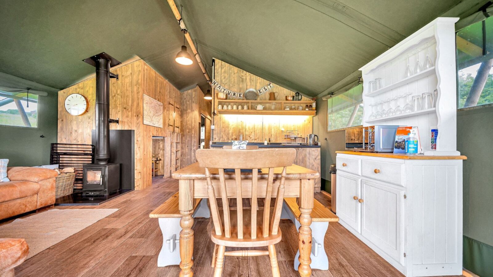 The cozy Nature’s Nest cabin interior boasts wood-paneled walls and a pitched ceiling. It features a wood-burning stove, a rustic dining table with chairs, shelves stocked with kitchenware, and a white hutch. Natural light filters in through windows, offering an idyllic glamping experience.