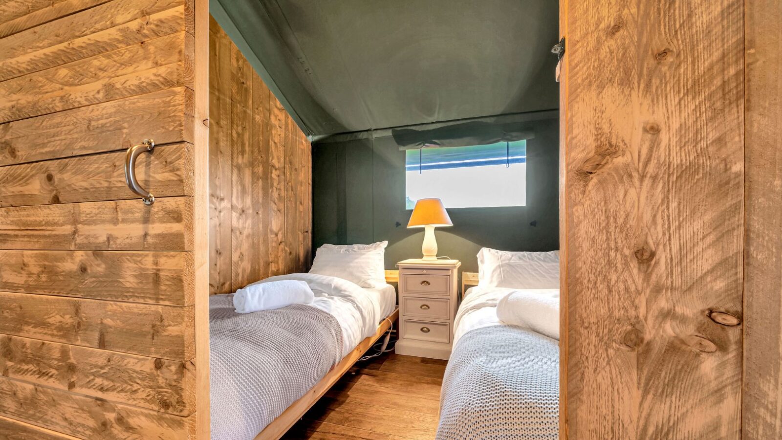 A cozy bedroom at Nature's Nest features two single beds with wooden frames and gray bedding. A small bedside table with a lamp sits between the beds, while rustic wooden walls add charm. A window above the table bathes the room in natural light, perfect for your glamping experience.