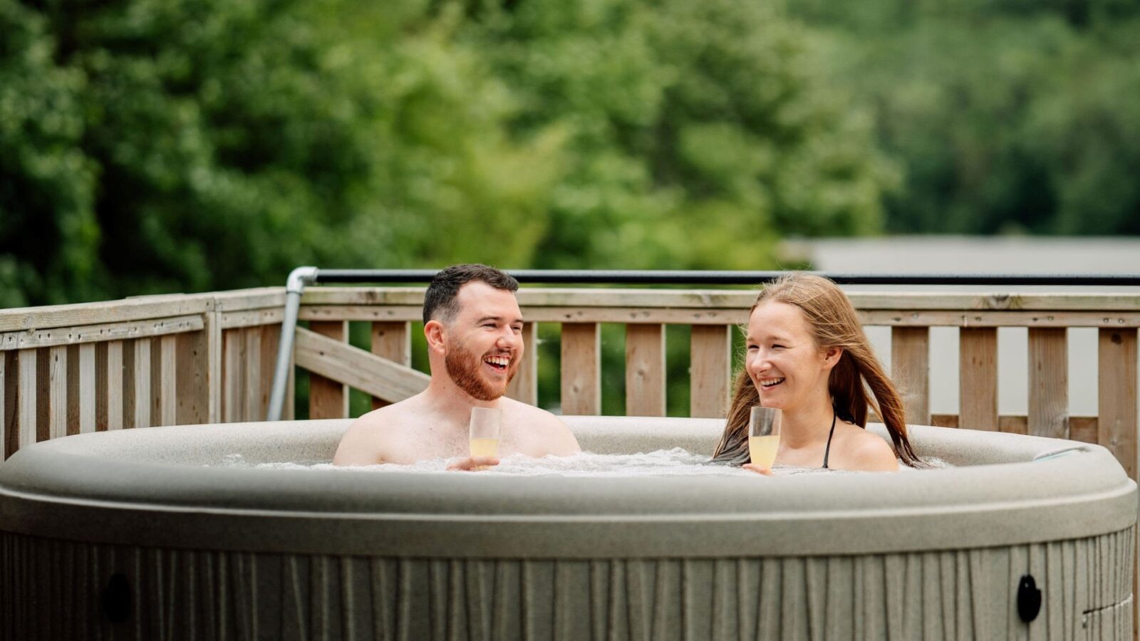 A man and a woman are enjoying a serene glamping experience in a hot tub on a wooden deck, surrounded by lush green trees. They smile and raise their glasses of champagne, embracing the tranquility of nature that envelops their cozy nest.