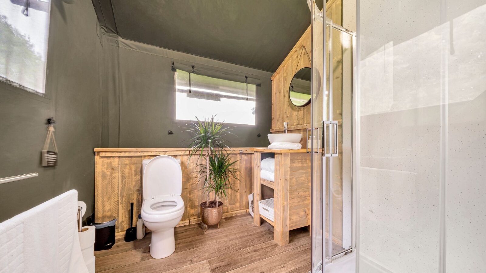 A modern bathroom in Nature's Nest with wooden accents features a toilet, a potted plant, and a walk-in shower with glass doors. A rustic wooden vanity with a round mirror and rolled towels is positioned against a wall with large windows, enhancing the glamping-inspired aesthetic.