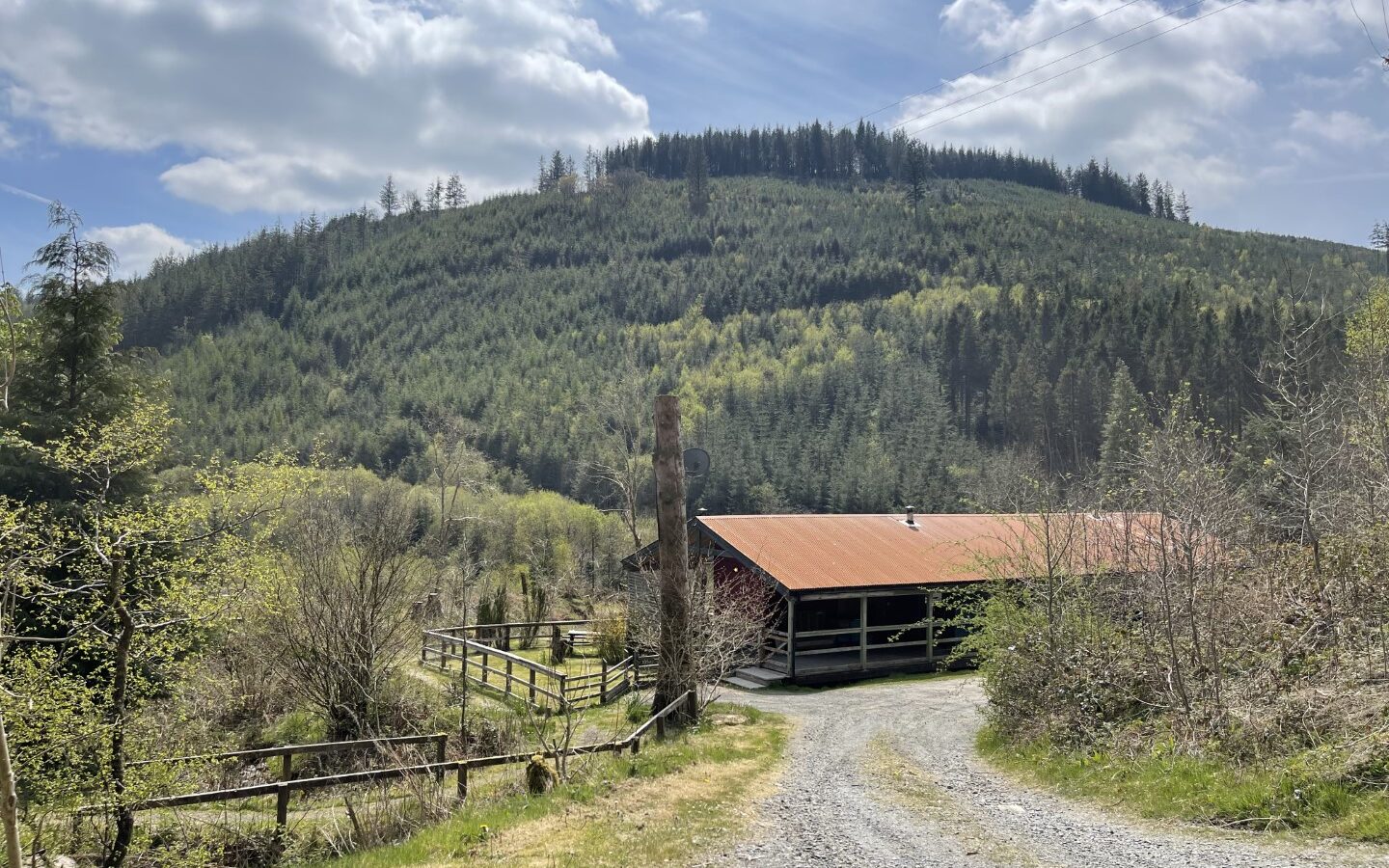 A gravel path leads to a barn with a red roof, nestled amid cottages and surrounded by trees and hills under a partly cloudy sky, in the heart of Trallwm.