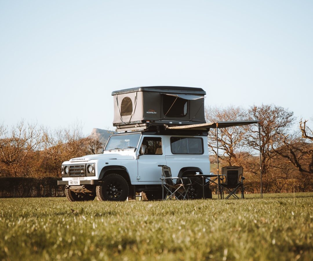 A white camper van with a roof tent, perfect for camping adventures, is parked on the grass, surrounded by folding chairs and trees in the background.