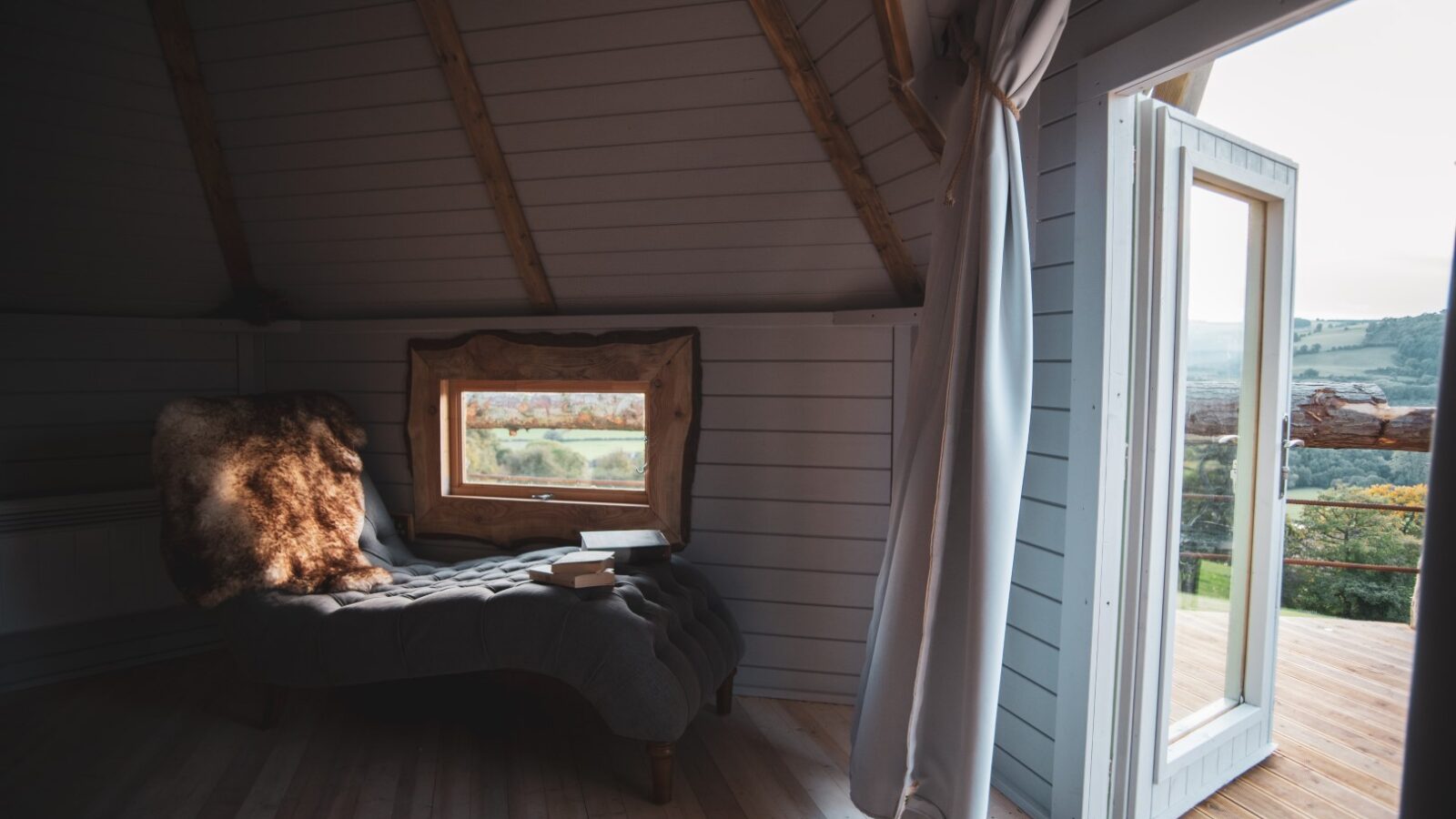 A cozy attic room with a lounged chair, small window, and an open door leading to a balcony where the landscape unfolds like a scene from dreams of dragons.