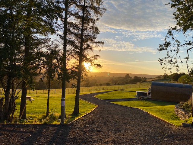 As the sun sets over a grassy landscape with trees, a picnic table invites relaxation near a small building in the distance. Meandering paths guide you through this idyllic scene, perfect for embracing the tranquility of farm glamping.