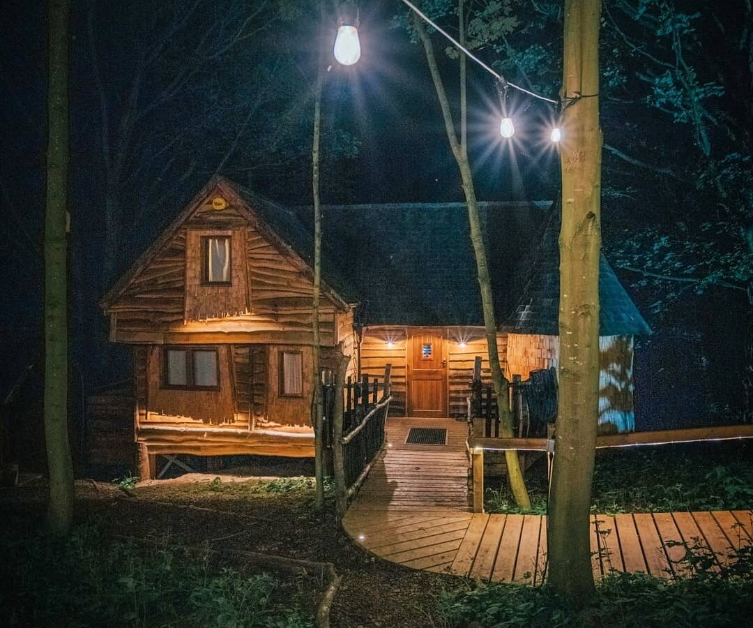 A cozy wooden cabin sits in a forest at night, illuminated by warm lights. A wooden footpath leads to the front door of the cabin, reminiscent of Rufus's Roost Treehouse at Baxby Manor. Hanging string lights are strung between trees above, adding a charming ambiance to the serene and picturesque setting.