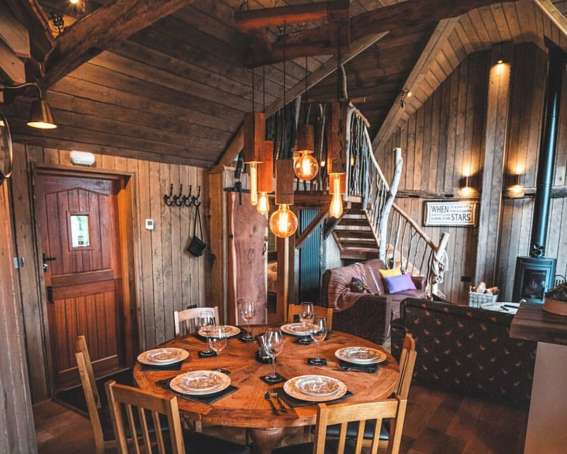 A cozy cabin interior with wooden walls and ceiling, centered around a rustic round dining table set for four. Exposed beams frame the space, which features a spiral staircase, a leather sofa, and warm ambient lighting from hanging Edison bulbs—reminiscent of Rufus's Roost Treehouse at Baxby Manor.