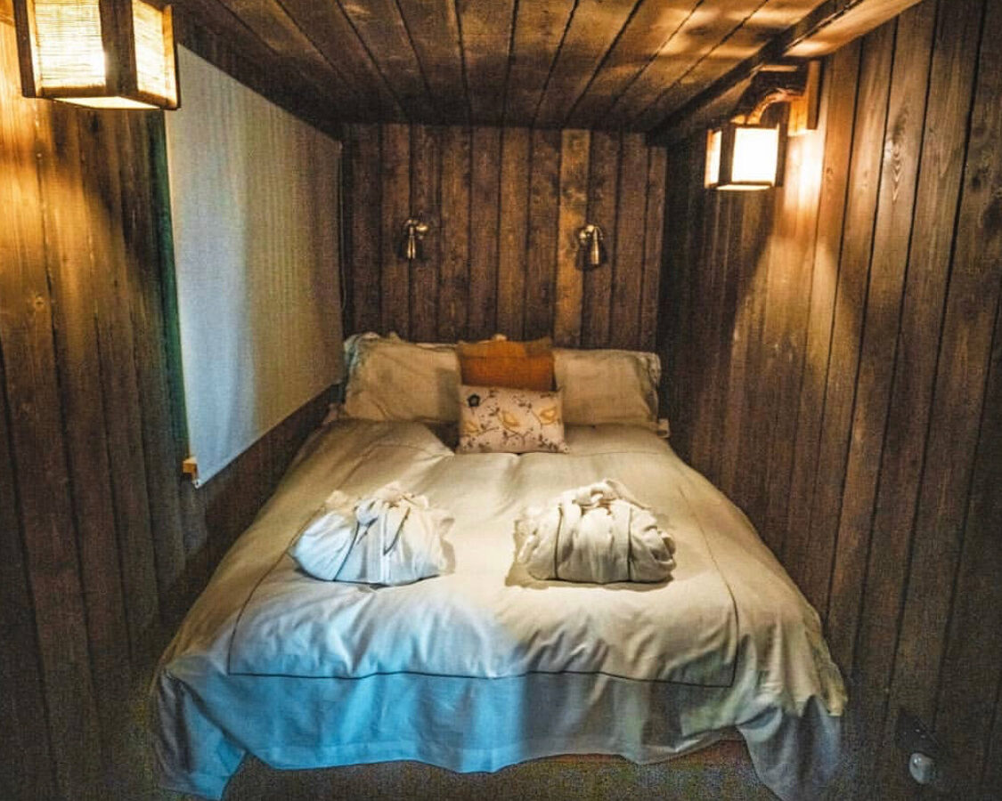 A cozy, intimate wooden cabin bedroom at Rufus's Roost Treehouse with dim lighting from wall-mounted lanterns. The bed is neatly made with white linens, two bathrobes, and decorative pillows. Warm wooden panels cover the walls and ceiling, creating a rustic ambiance reminiscent of Baxby Manor.