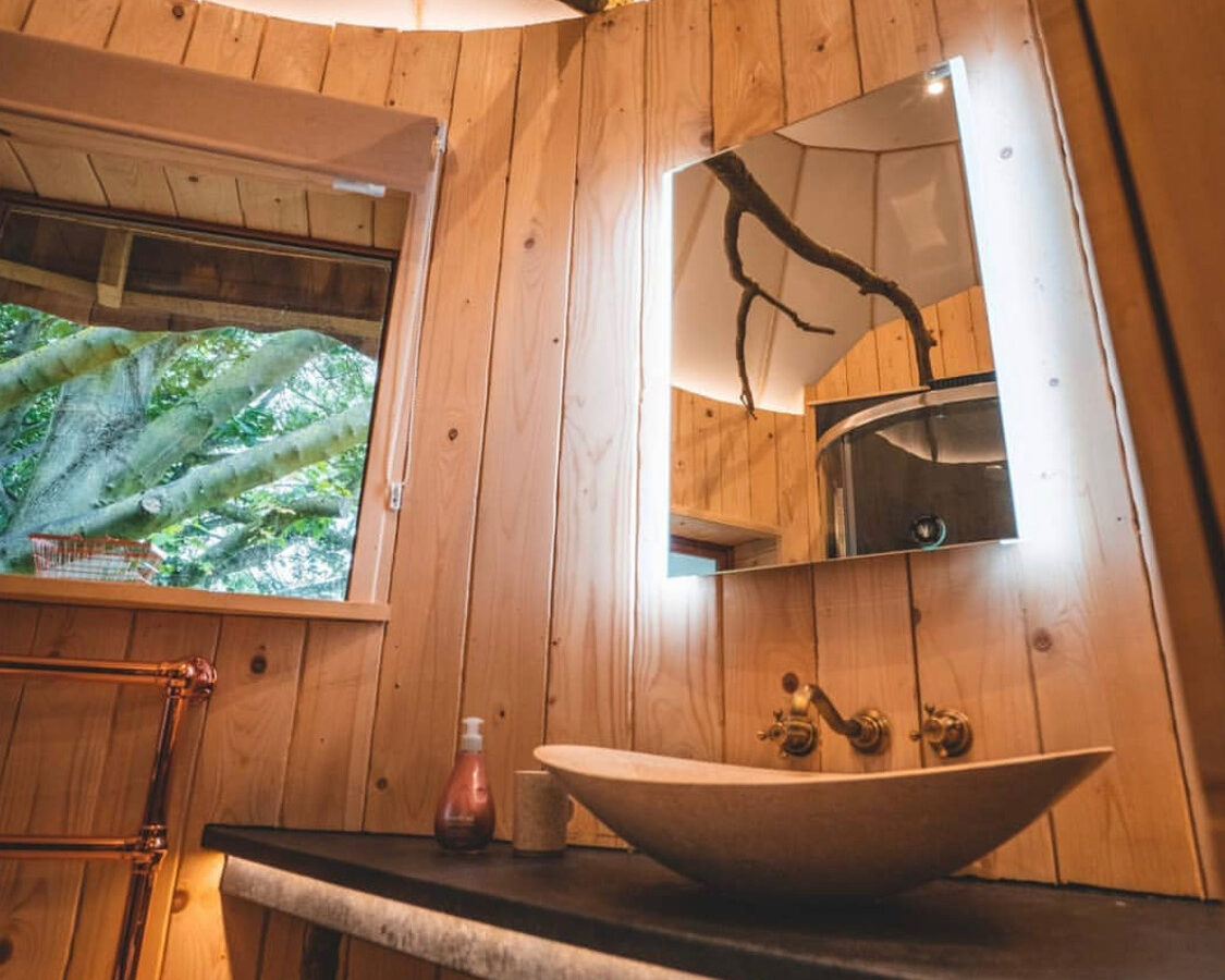 A cozy, rustic bathroom with light wood-paneled walls reminiscent of Baxby Manor. A circular mirror with soft backlighting hangs above a stone vessel sink on a dark wooden countertop. A window on the side lets in natural light and a view of green foliage outside, enhancing the nature-inspired decor.