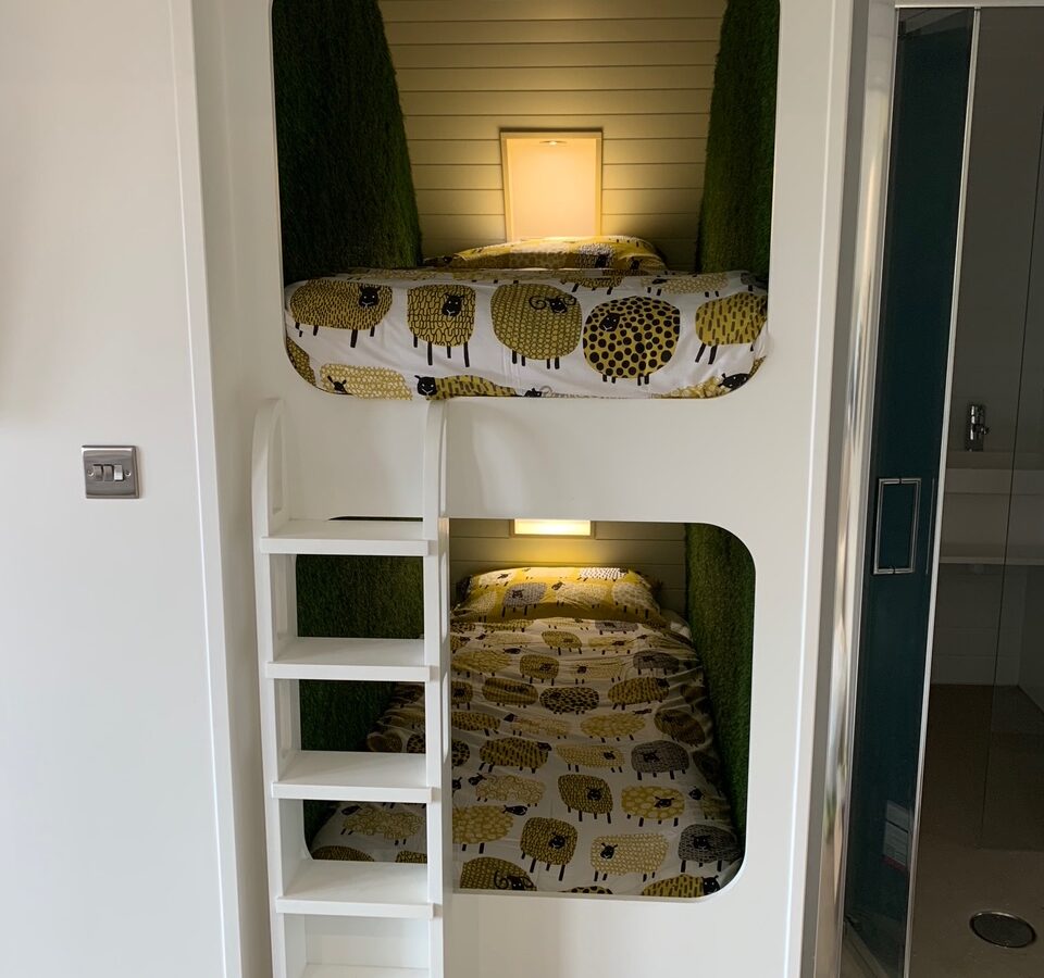 Two built-in bunk beds with white frames and sheep-themed bedding capture the essence of glamping at Great House Farm. A small ladder provides easy access to the top bunk, blending rustic charm with modern comfort.