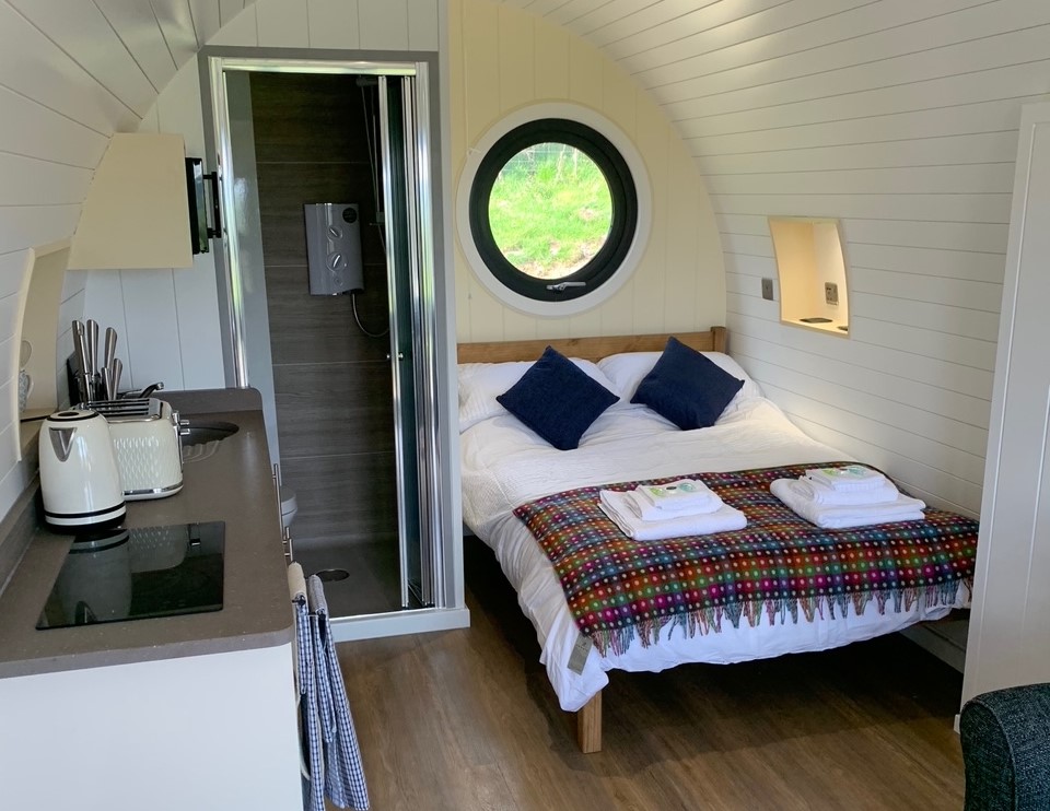 Compact modern cabin interior with a bed, kitchenette, and small bathroom. A circular window above the bed offers serene farm glamping views; a colorful blanket adds charm to your stay. Experience the tranquility of Great House Farm right from your cozy retreat.