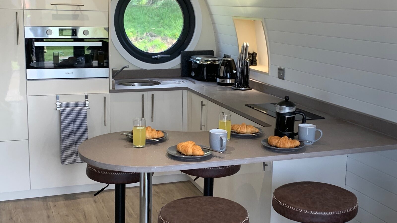 A modern kitchen with a round window and breakfast bar evokes a cozy glamping vibe. Plates with croissants, coffee mugs, and juices adorn the counter, inviting you to enjoy the comforts of house farm living. Stools line up neatly, ready for a leisurely morning meal.