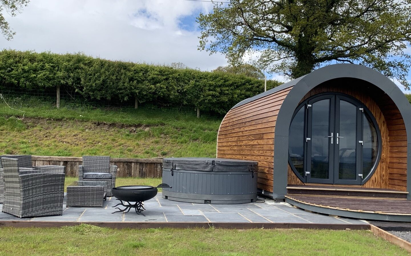 Experience glamping at Great House Farm in this charming wooden pod cabin. With large glass doors, outdoor furniture, a hot tub, and a tree framing the background, it's your perfect escape into nature's embrace.