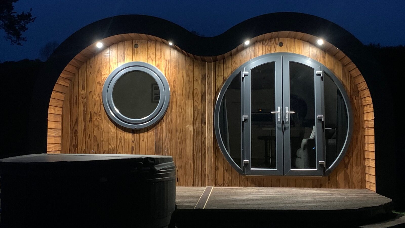 A modern wooden cabin with round windows and doors, illuminated at night, offers a cozy glamping experience. Nestled on the Great House Farm grounds, it features a hot tub on a grassy area, perfect for relaxation under the stars.