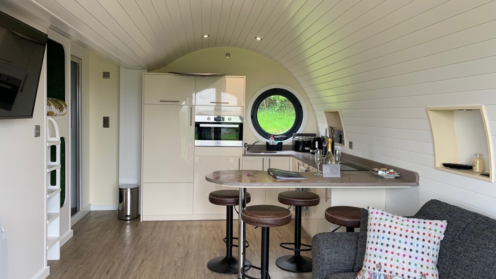 Modern tiny home interior with white walls, a kitchenette, round window, bar stools, and a gray sofa with a colorful pillow. Perfect for those seeking the cozy charm of glamping at Great House Farm.