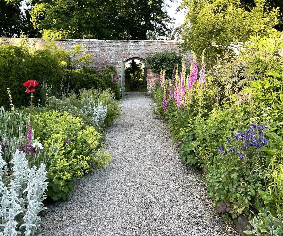 Gravel path through a lush garden with colorful flowers and greenery, leading to a brick archway, creating an enchanting retreat.