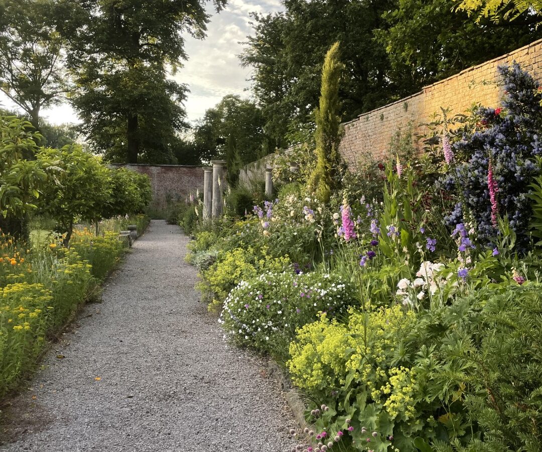 A gravel path bordered by lush green plants and colorful flowers leads to a brick wall with tall trees in the background, offering a sense of relaxation akin to a serene laundry retreat.