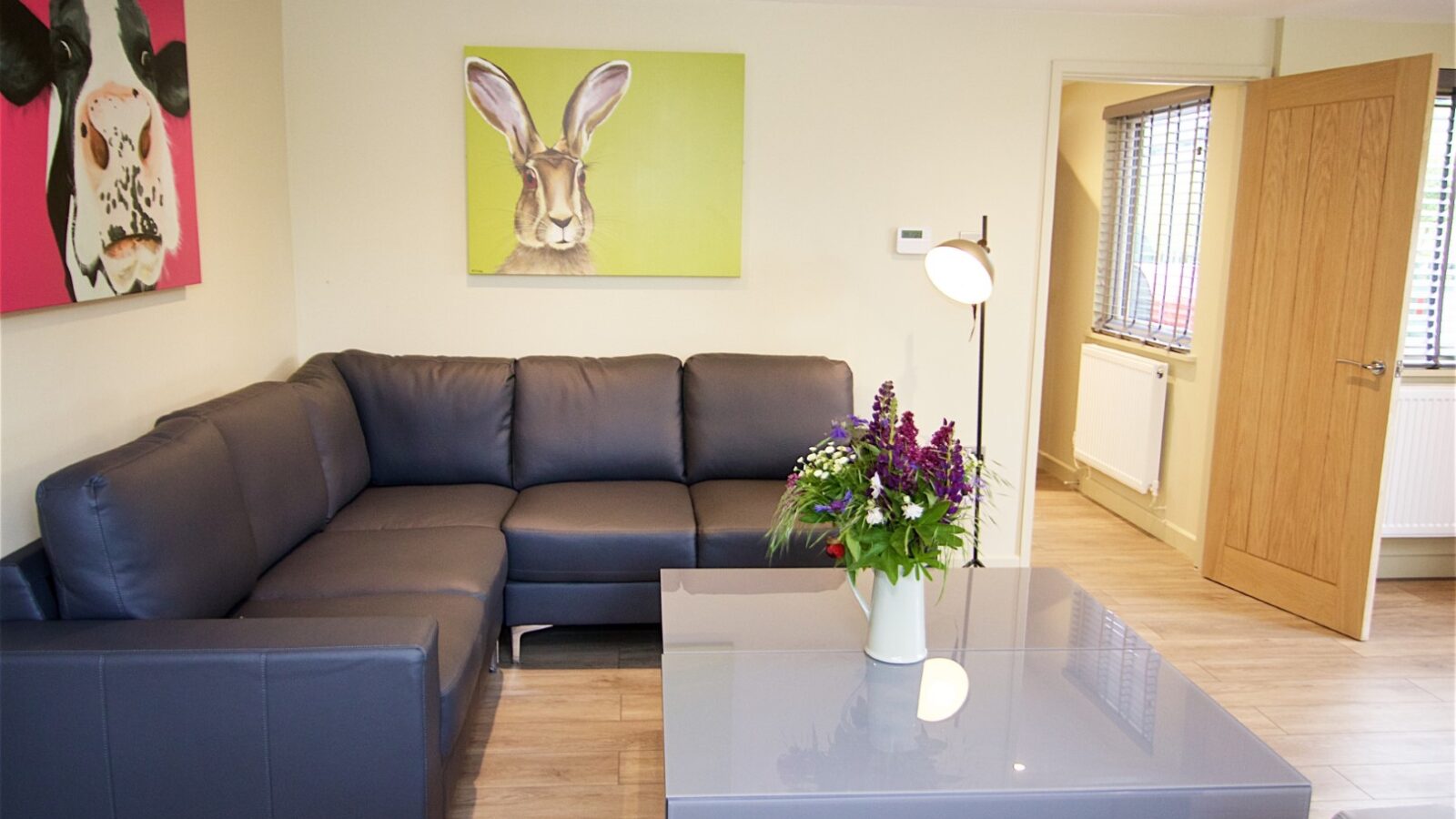 Modern living room with a gray sectional sofa reminiscent of a forest retreat, paintings of a cow and rabbit adding charm, and a large coffee table with a floral arrangement. This tranquil space feels like Jess’ Tree Bach: Treehouse, offering the perfect nature stay indoors.