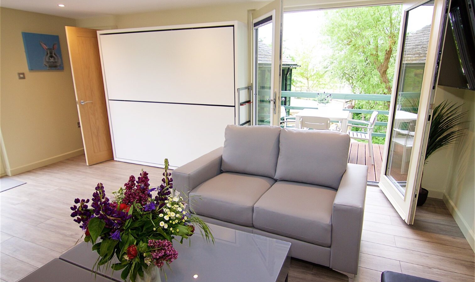 Bright living room in Jess’ Tree Bach: Treehouse, featuring a gray sofa and wall bed. Fresh flowers on a table complement the open double doors leading to a balcony with outdoor seating, creating a serene nature retreat.