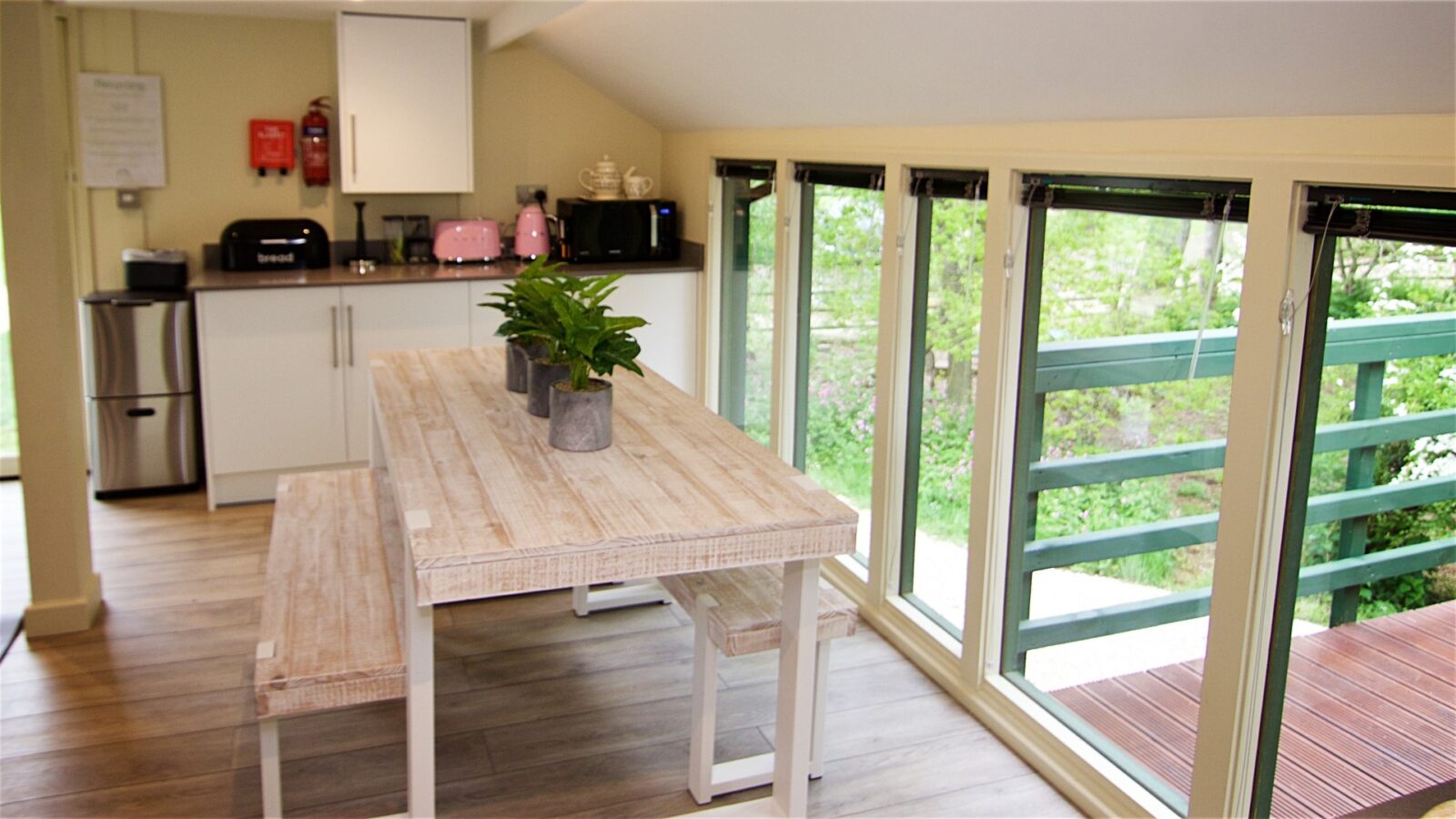 Bright kitchen with a wooden table, benches, and potted plants, seamlessly merging nature with comfort. Large windows invite natural light into this treehouse haven. Appliances include a toaster and kettle for your morning rituals. Welcome to Jess' Tree sanctuary.