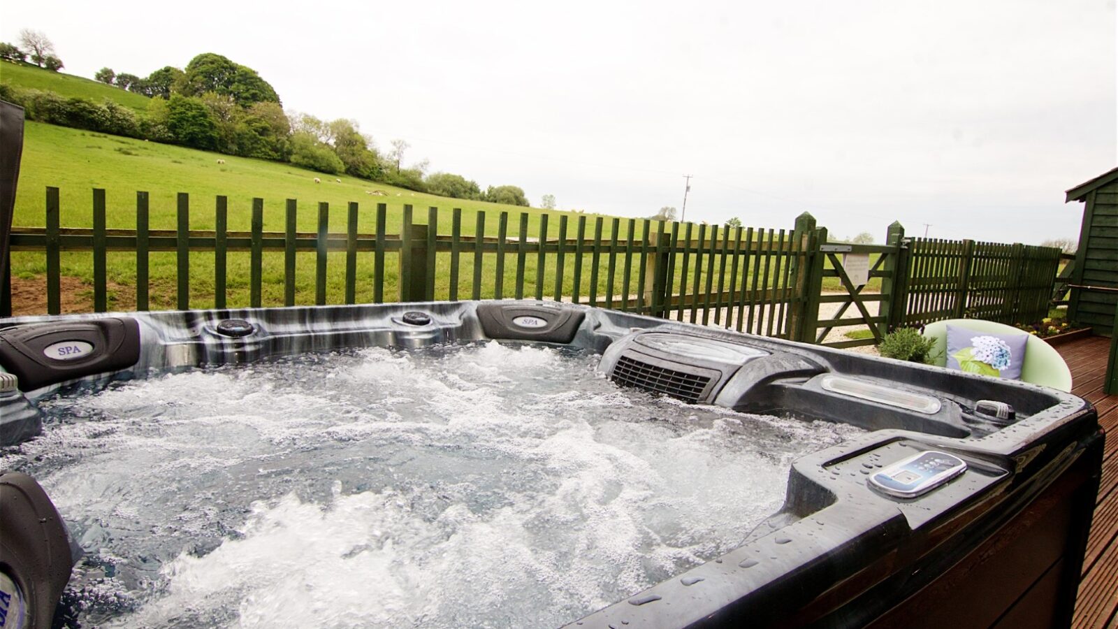 Experience a rustic getaway with a hot tub bubbling on the deck of Jess’ Tree Bach: Tree House Accommodation, overlooking a fenced grassy field and serene hills in the background.