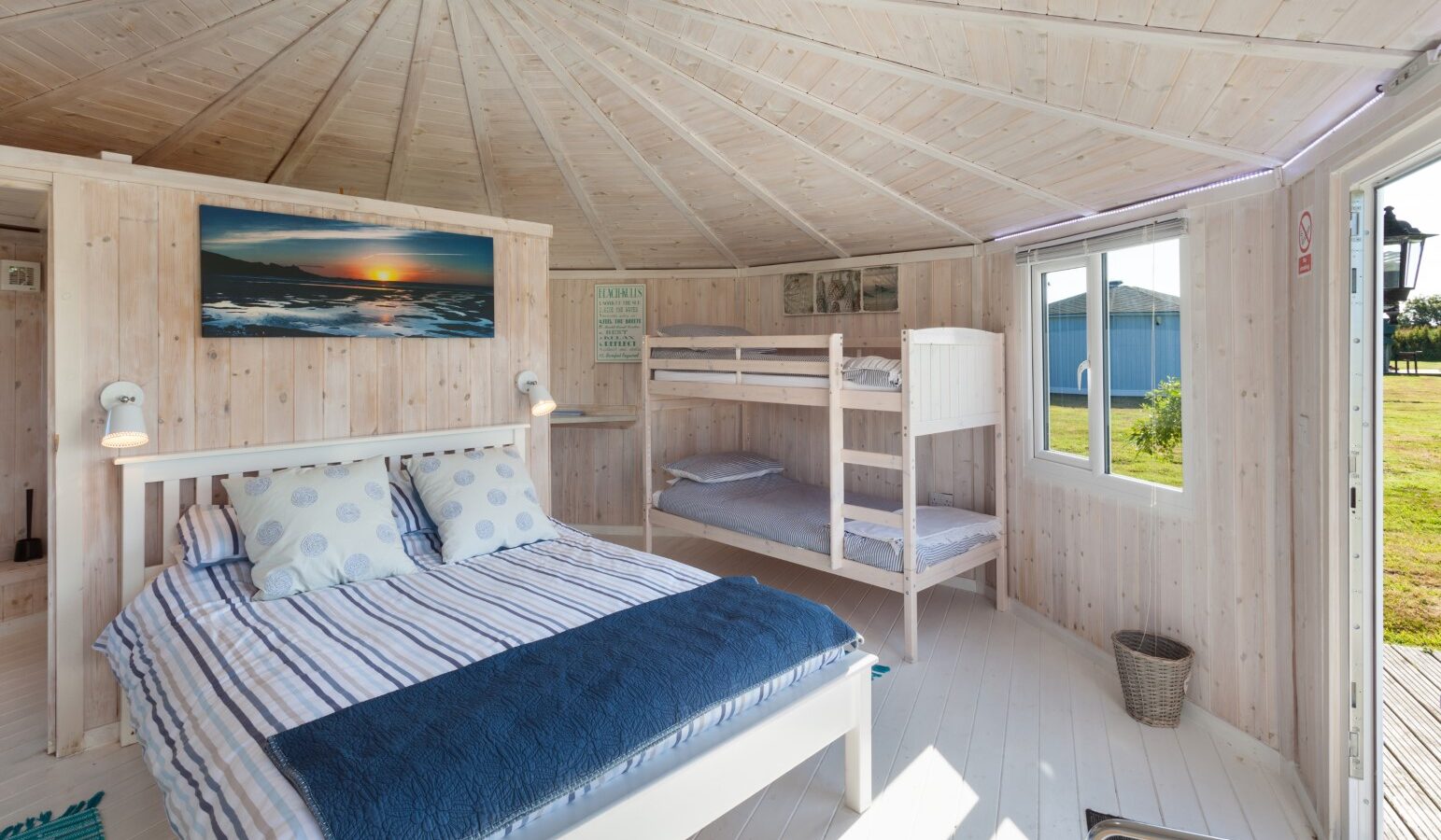 Cozy coastal cabin bedroom with a double bed, bunk beds, a landscape painting, and a large window overlooking the grassy expanse outside.