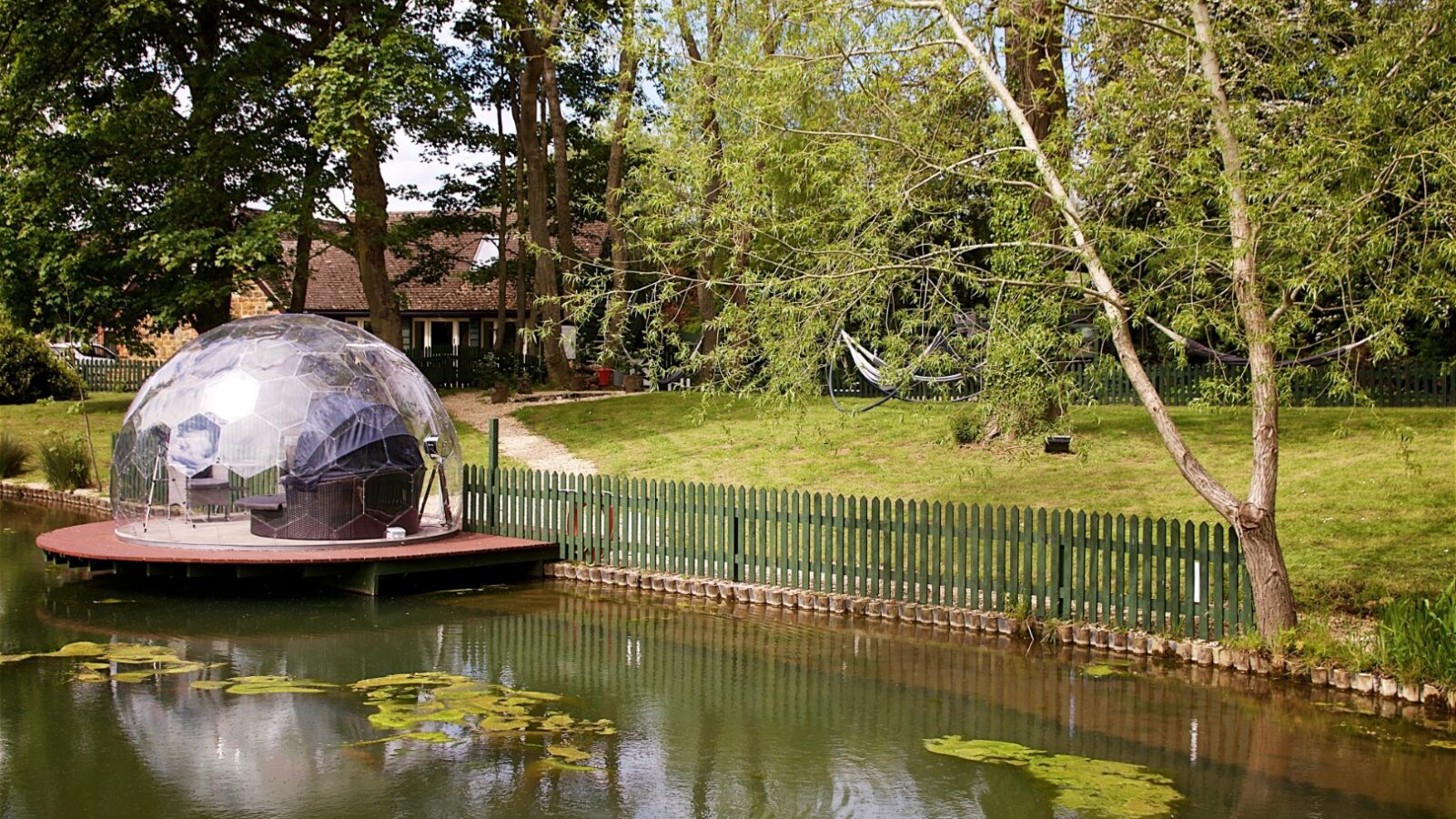 A dome tent beside a tranquil pond in Jess’ Tree Bach garden setting, with a cozy house and trees gracing the background.