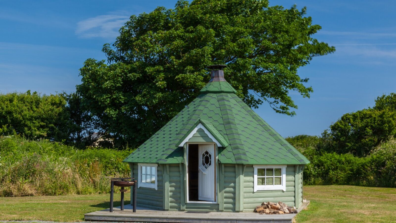 A small green hexagonal coastal cabin sits on a grassy lawn with trees in the background, featuring a single door and window.