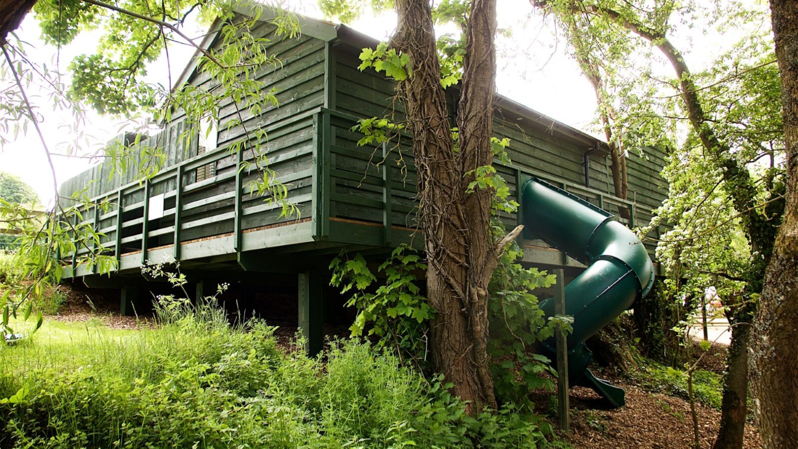 Nestled amidst lush trees and greenery, the green wooden treehouse, affectionately called Jess's retreat, features a slide that offers endless fun in its enchanting outdoor setting.