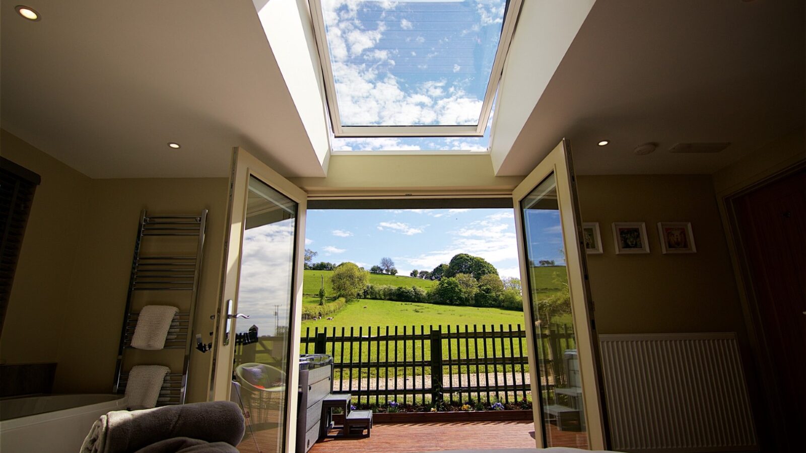 The bedroom view, reminiscent of a serene Bach composition, features open doors leading to a deck. Beyond lies a scenic landscape with lush green fields and a vast blue sky, framed perfectly through the skylight.