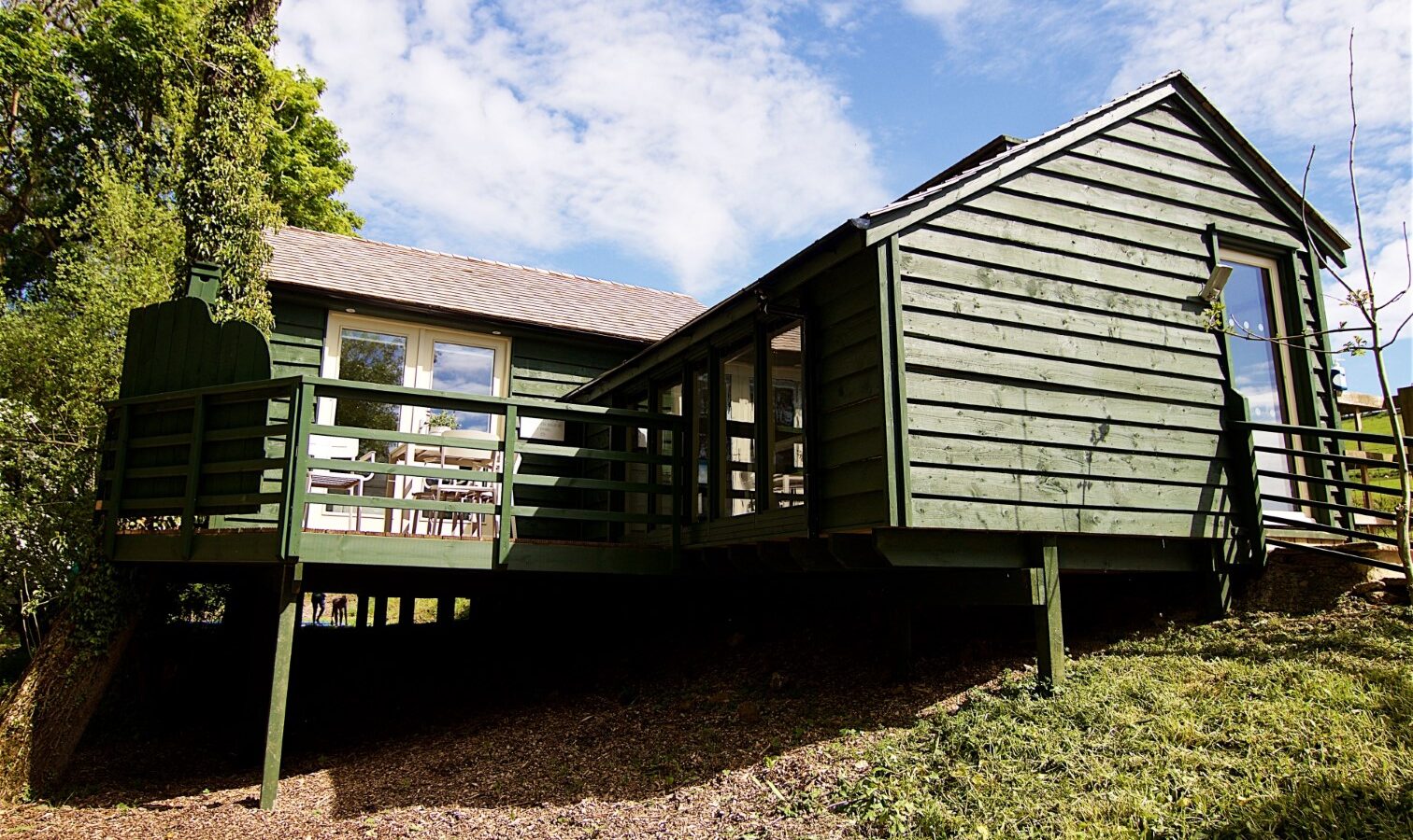 A charming green wooden cabin with large windows and a deck, nestled like a treehouse among the trees. It offers a serene escape with its sloped grassy area under a brilliant blue sky.