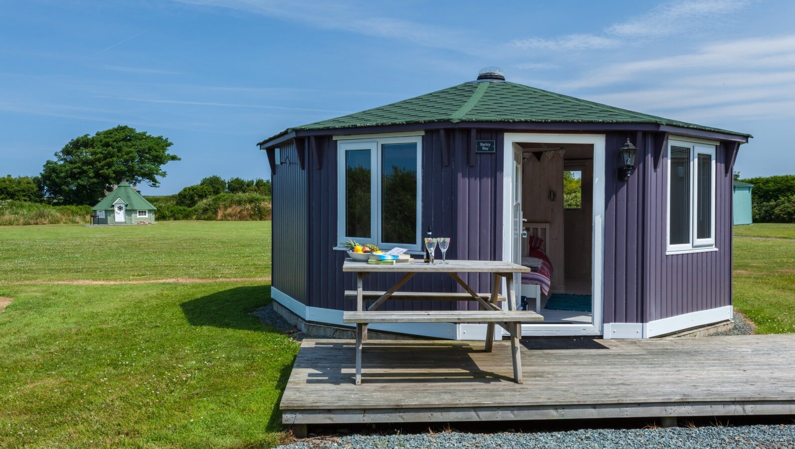 A purple yurt with white trim stands on a grassy field like a cozy cabin retreat, featuring a picnic table adorned with a bowl and glasses, all under the expansive blue sky just beyond the coastal horizon.