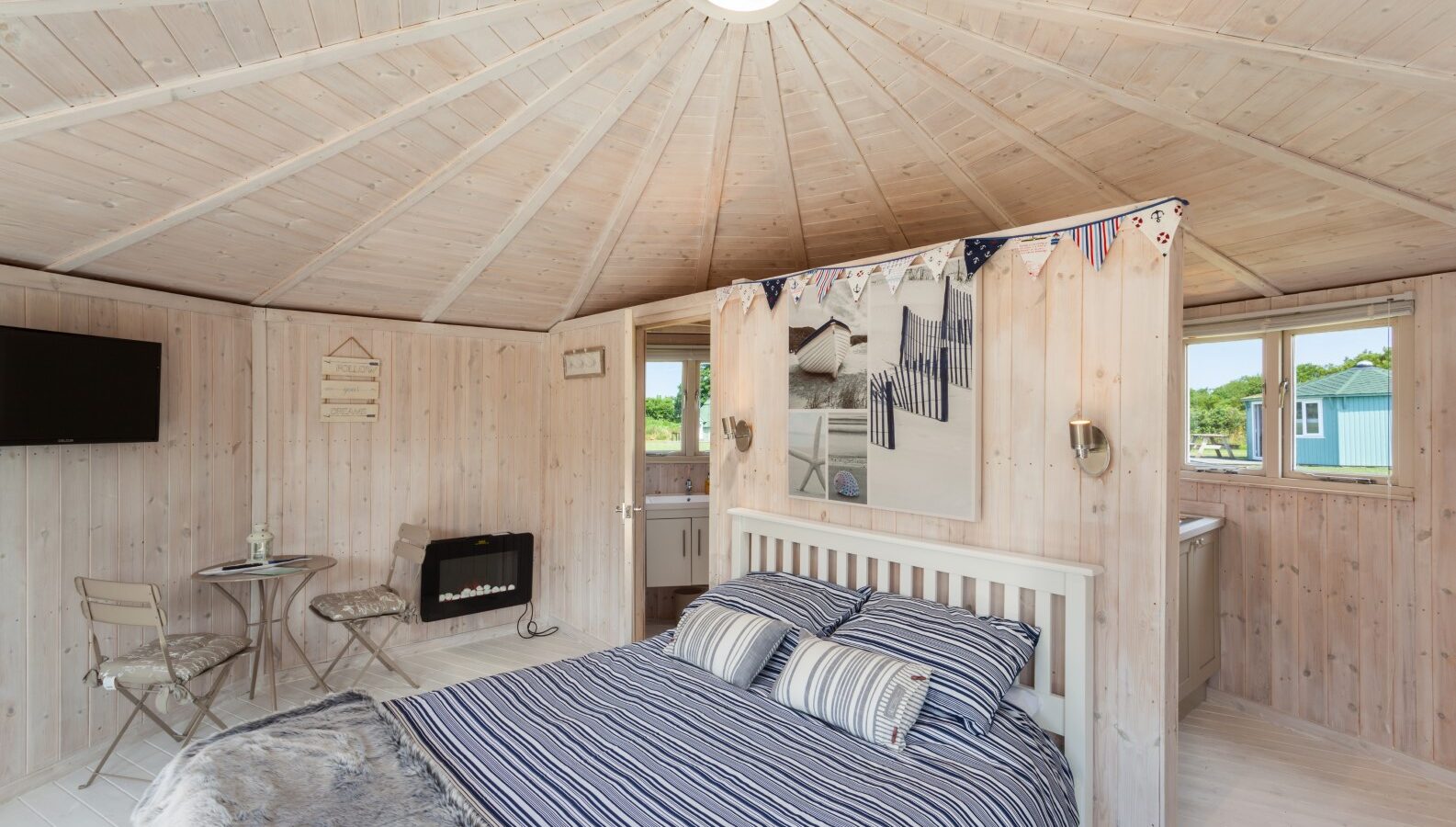 Cozy wooden cabin bedroom with a double bed, striped bedding, and a TV, complemented by a small table and chairs. The kitchenette in the background completes this idyllic coastal retreat.