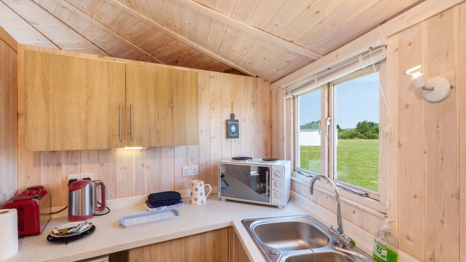 A cozy kitchenette with wooden walls, counter, sink, microwave, and tea kettle evokes the charm of coastal cabins. The window offers a grassy outdoor view.