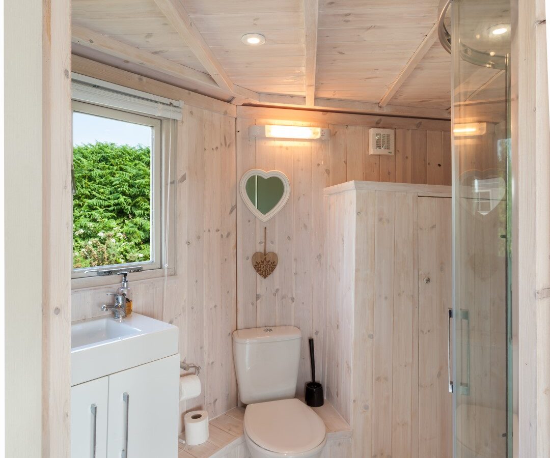 Charming small bathroom with wood paneling evokes the rustic elegance of coastal cabins. It features a toilet, sink, curved glass shower, and heart-shaped mirror. A window offers a tranquil view of lush greenery.