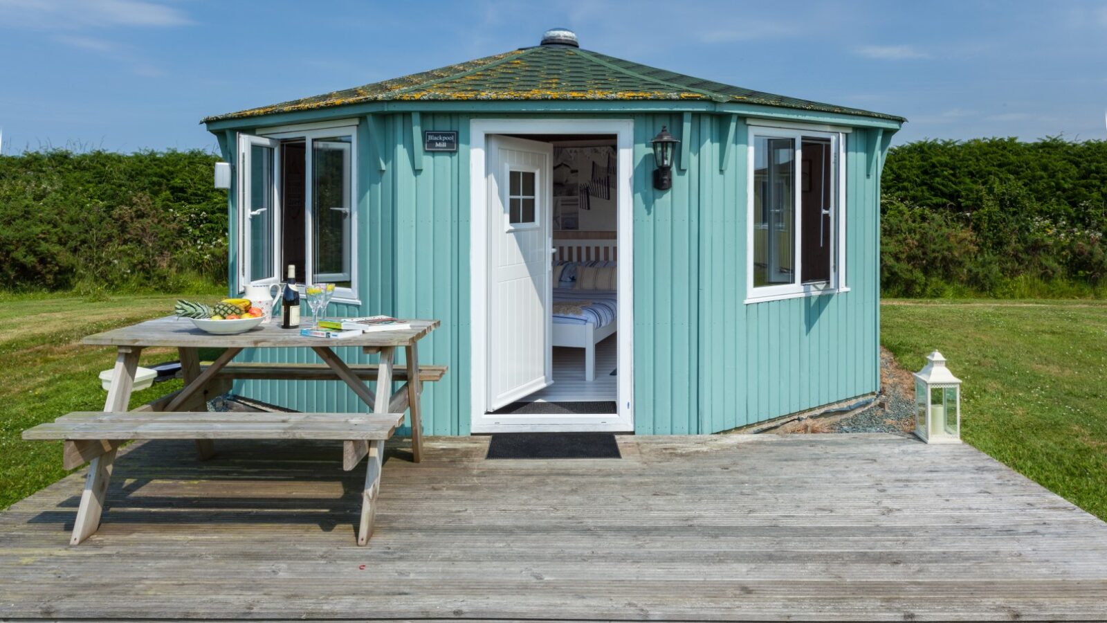 A charming, small round turquoise coastal cabin with a white door sits on a wooden deck. In the foreground, a picnic table is set with food and drinks, inviting you to relax and savor the ocean breeze.