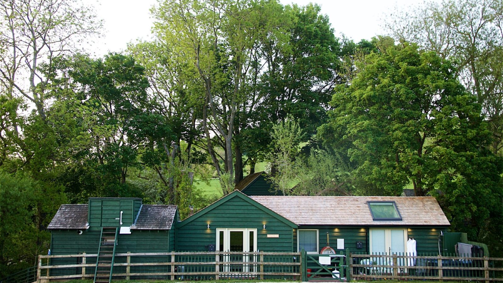 Jess’ Tree Bach: This green wooden cottage, nestled among the trees, serves as a perfect nature escape. With a fence in the foreground and a clear sky above, it's an idyllic holiday rental that promises tranquility and rejuvenation amidst nature.