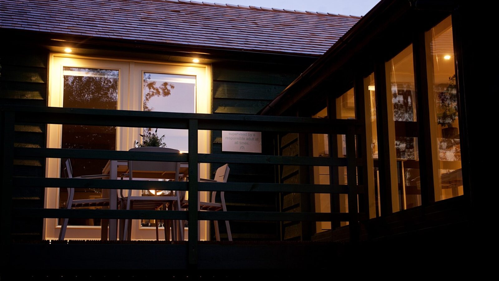 A wooden cabin nestled among the trees, with illuminated windows and a deck featuring a table and chairs, echoes the tranquil notes of Bach as dusk softly descends.