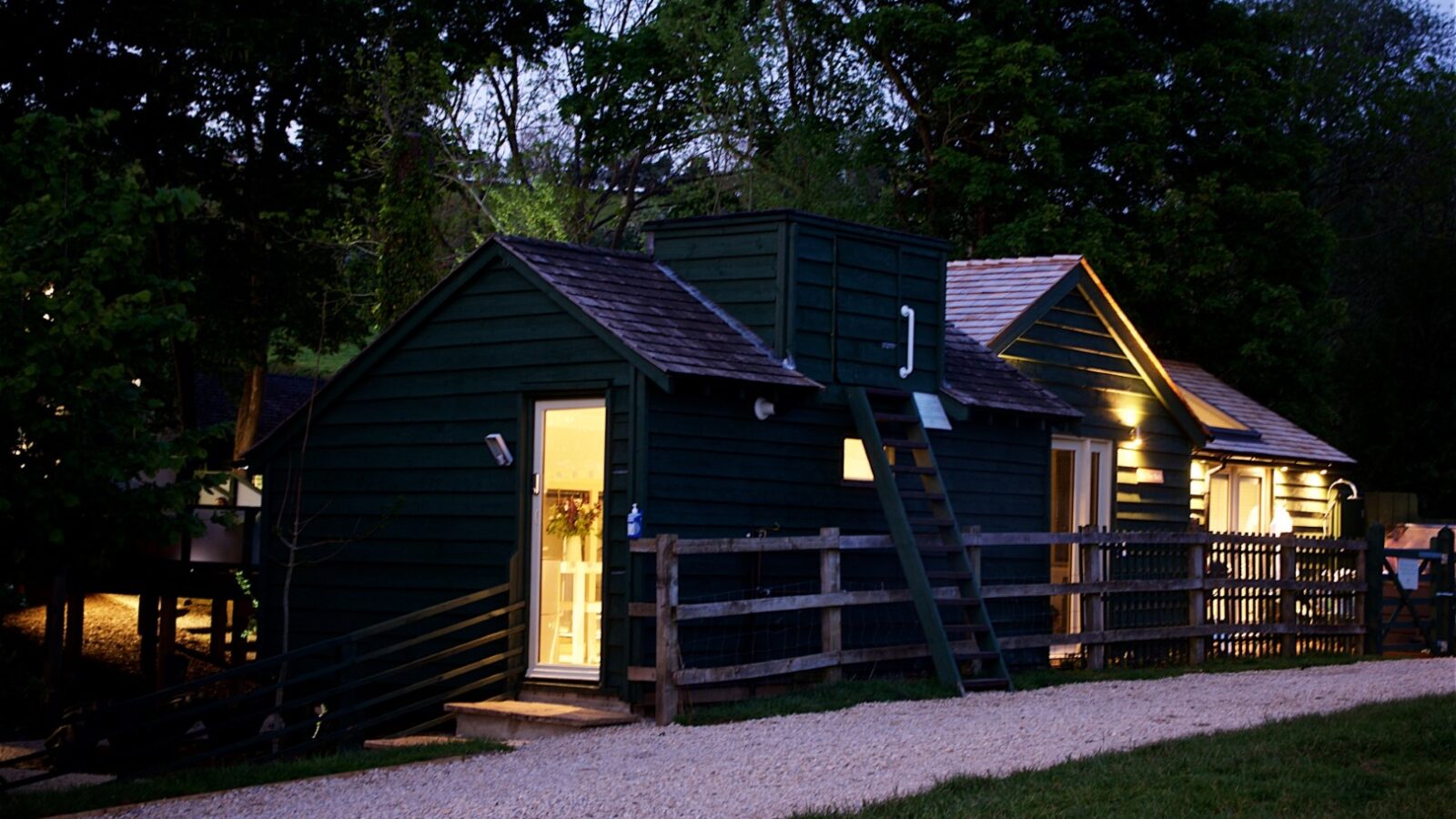 A small green wooden cabin, perfect for a serene forest retreat, stands with its lit windows surrounded by trees at dusk. A gravel path and wooden fence frame this idyllic cabin getaway.