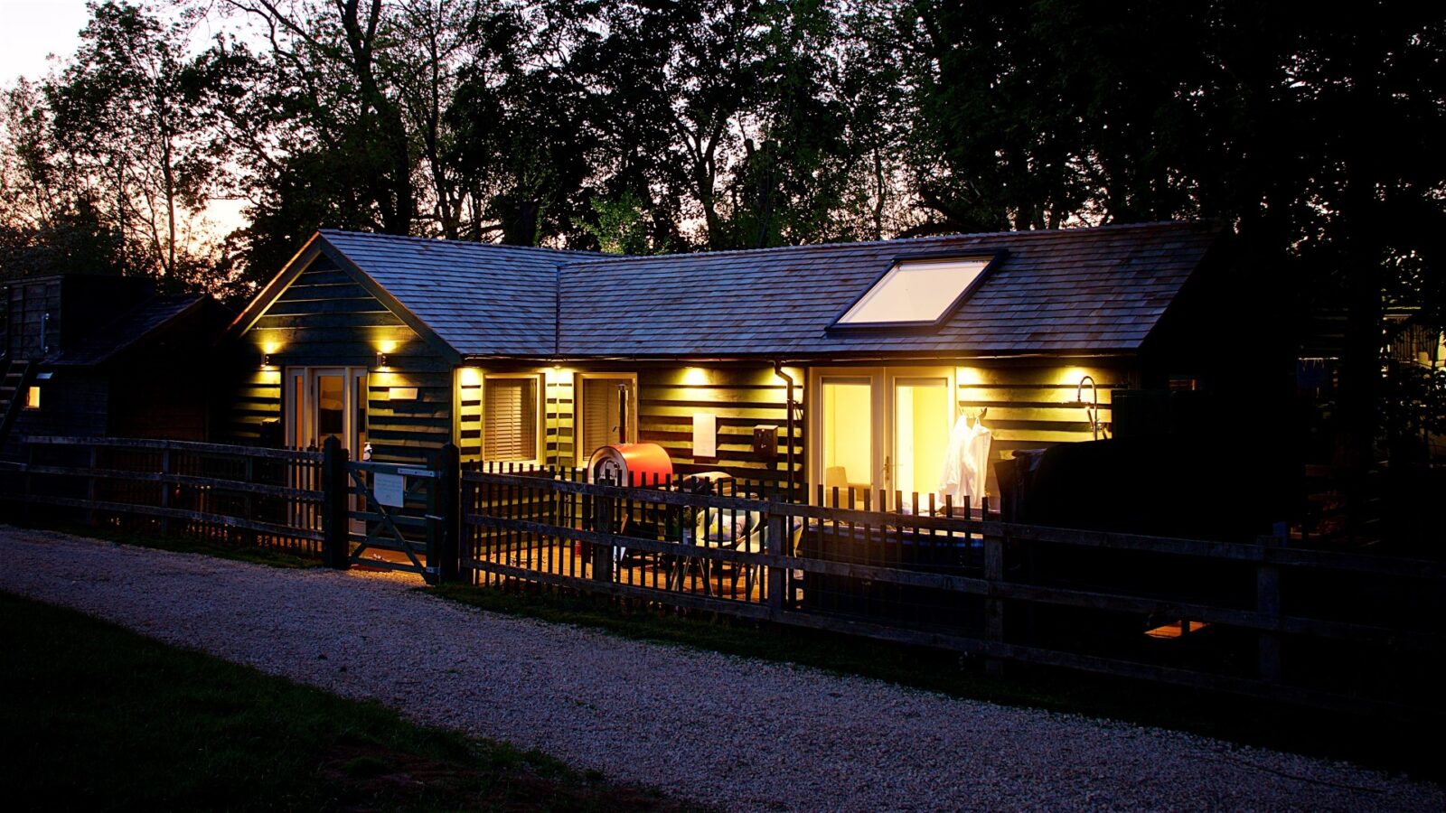 A cozy, illuminated cabin known as Jess’ Tree Bach with a fenced yard at dusk is surrounded by trees, while a gravel path winds invitingly in the foreground.