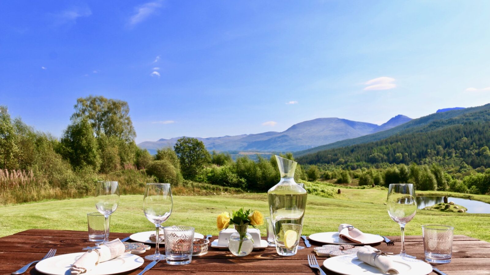 A charming outdoor dining setup on a wooden table with plates, glasses, and a carafe sits like a scene from a Frances sketch, beautifully framed against the backdrop of a scenic mountain landscape.