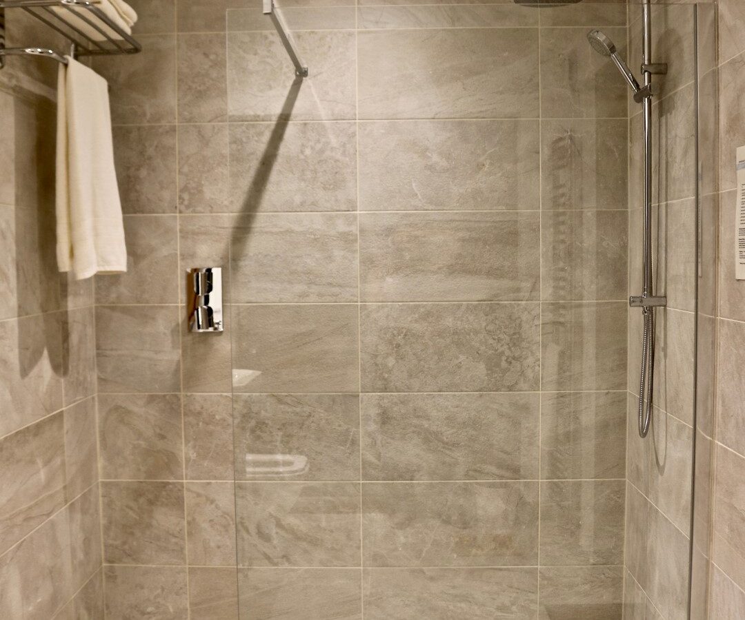 Modern bathroom shower with gray tiled walls, a glass partition, and a rain showerhead. A towel rack with folded towels adds a touch of simplicity, inviting Frances to sketch the serene setting on her sketch pad as an artful study in tranquility.