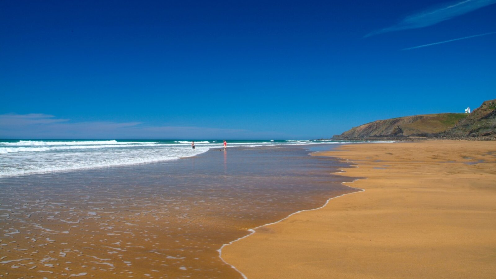 Wide sandy beach with gentle waves and a clear blue sky. Distant figures enjoy the coastal waters while charming cabins dot the horizon near the cliff.