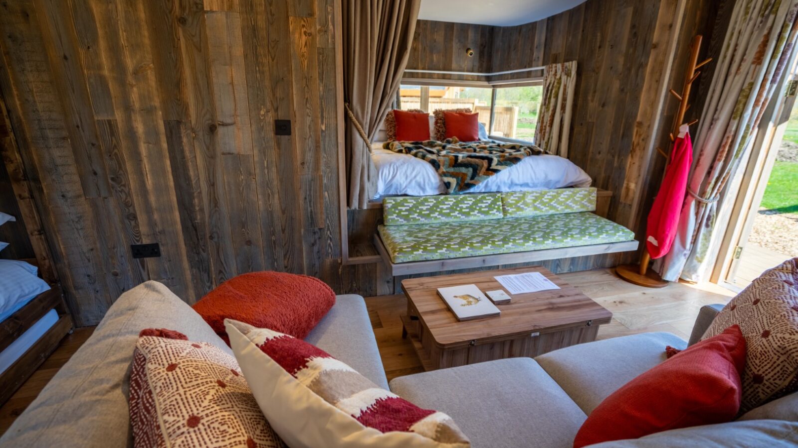 A cozy, rustic cabin interior at Baxby Manor, featuring wooden walls with a comfortable bed tucked behind a curtain, a sofa adorned with red and white pillows, a wooden coffee table, and a window showcasing lush greenery. The warmly lit Kabinas creates a serene and inviting atmosphere.