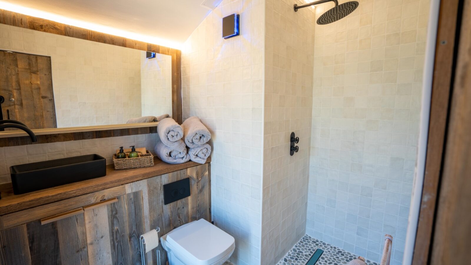 A modern bathroom at Baxby Manor features a walk-in shower with a black rain showerhead, white tile walls, and a pebble-tiled floor. The wooden vanity with its black sink and large mirror above adds style, while folded towels and toiletries are neatly placed on the wooden shelf.