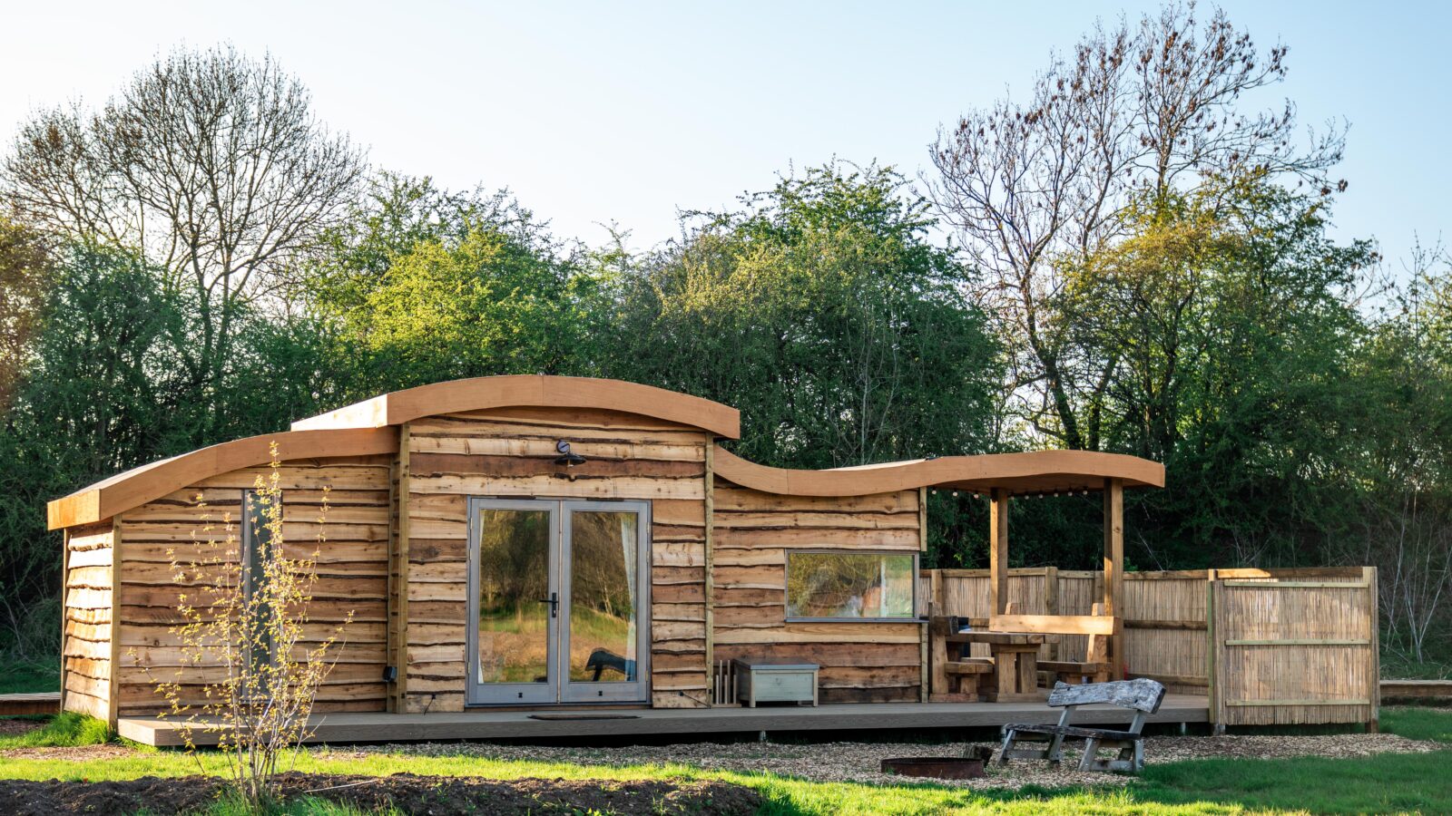 A modern, eco-friendly cabin by Kabinas with wavy, natural wooden siding is set in a lush, green environment. It features large glass doors, a small patio with seating, and a wooden fence. Trees in the background hint at a peaceful, serene setting under a clear sky near Baxby Manor.