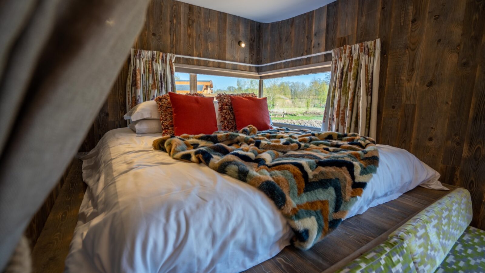 A cozy bedroom at Baxby Manor features wood-paneled walls and a large bed adorned with a fluffy, multi-colored chevron blanket and two red pillows. The room has large windows with curtains, offering a serene view of greenery and a distant building outside.