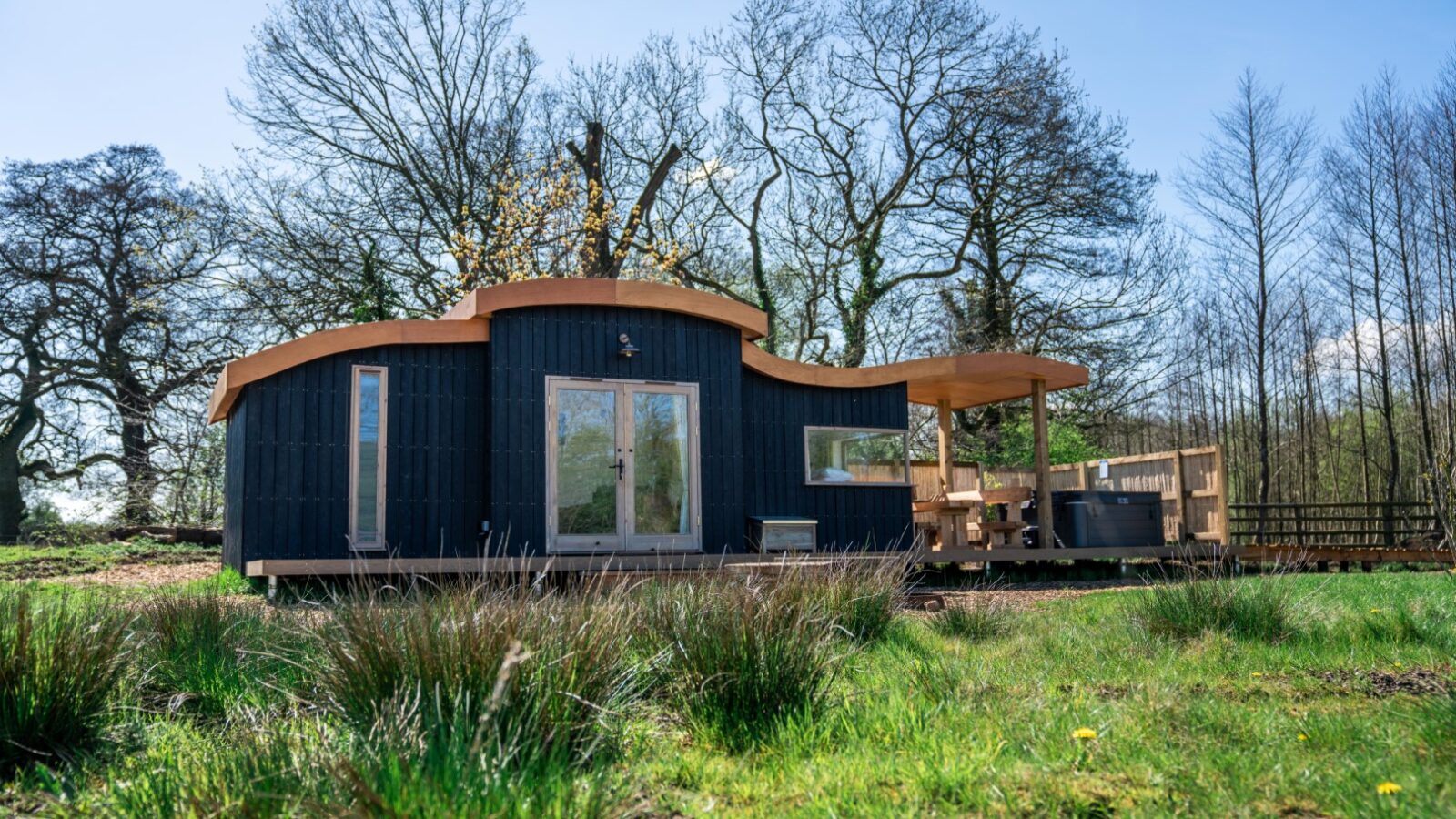 A modern, eco-friendly tiny house with a curved roof sits in a bucolic woodland setting at Baxby Manor. Featuring large glass doors, a wooden deck with seating, and surrounded by tall trees and lush grass under a clear blue sky, this Kabinas retreat is perfect for nature lovers.